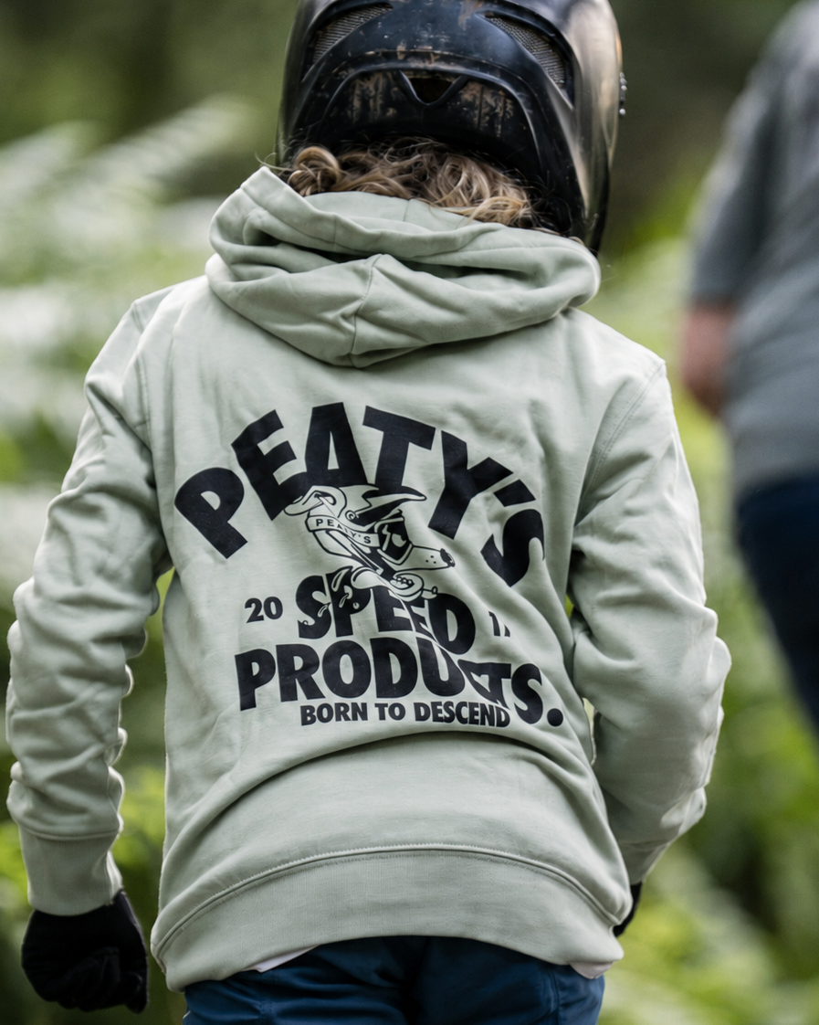 A child viewed from behind wearing a light sage green hoodie with a large black graphic on the back that reads 'PEATY'S 20 SPEED PRODUCTS BORN TO DESCEND,' along with a black helmet and blue pants, in an outdoor forest setting.