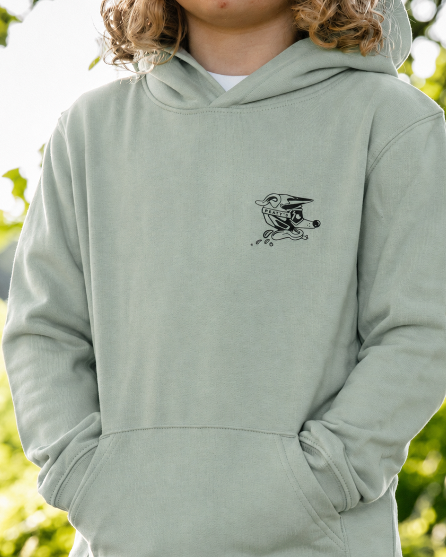 A child wearing a sage green pullover hoodie with a small black bird graphic on the left chest, photographed outdoors against a blurred green foliage background.