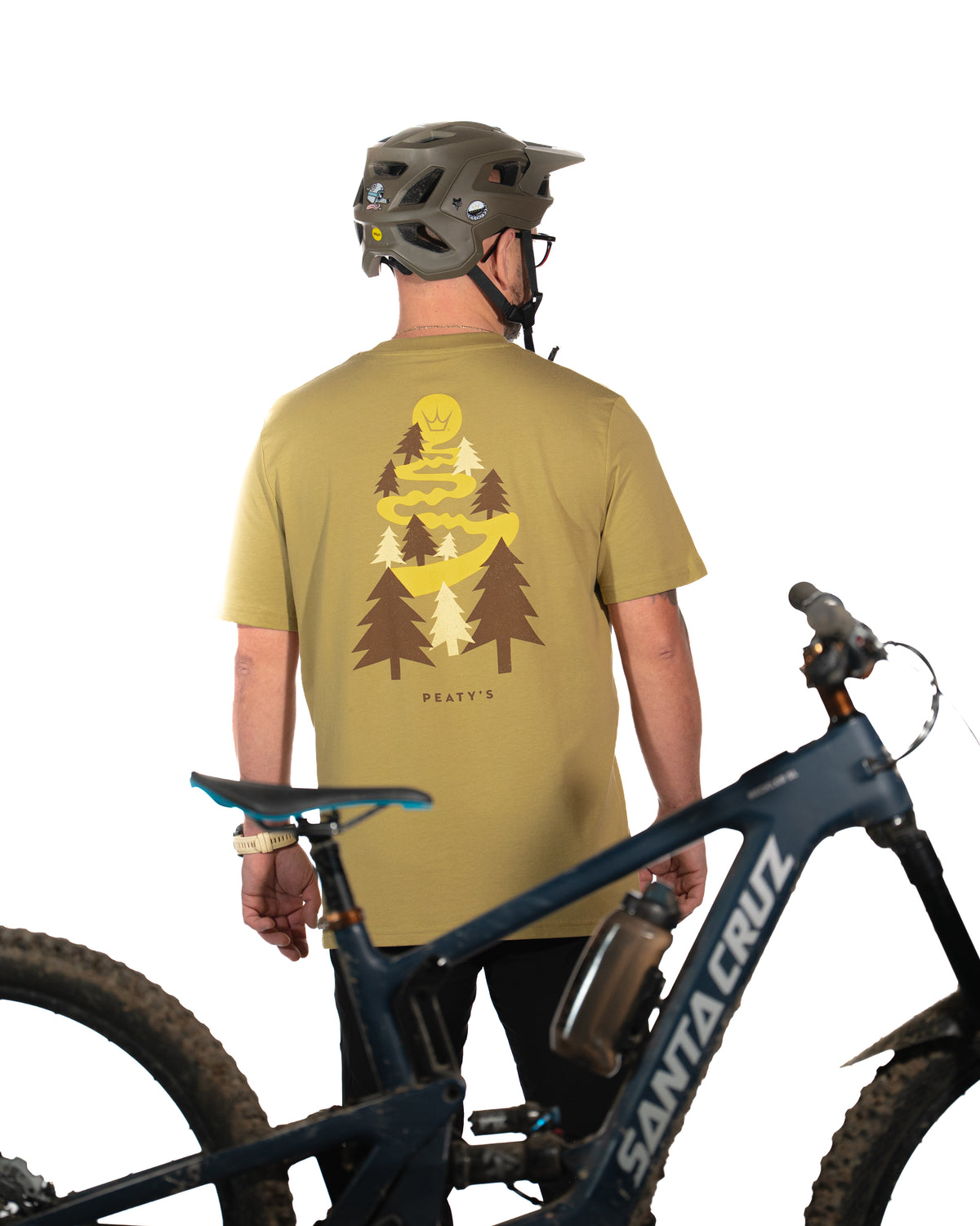 Rider wearing the Olive Peaty's Dusk to Dawn T-shirt beside a mountain bike, showing the illustrated back print