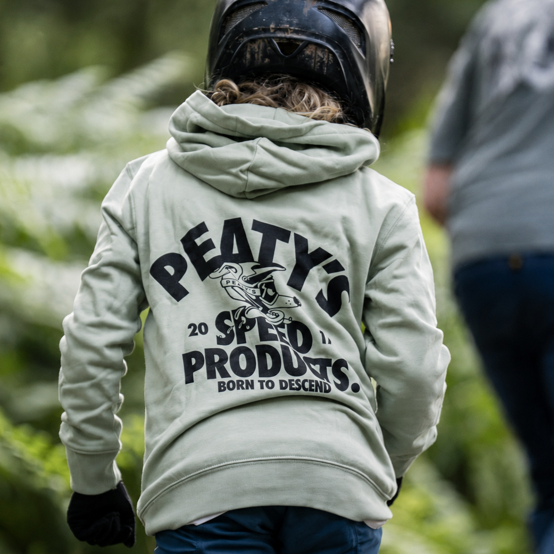 A child viewed from behind wearing a light sage green hoodie with a large black graphic on the back that reads 'PEATY'S 20 SPEED PRODUCTS BORN TO DESCEND,' along with a black helmet and blue pants, in an outdoor forest setting.