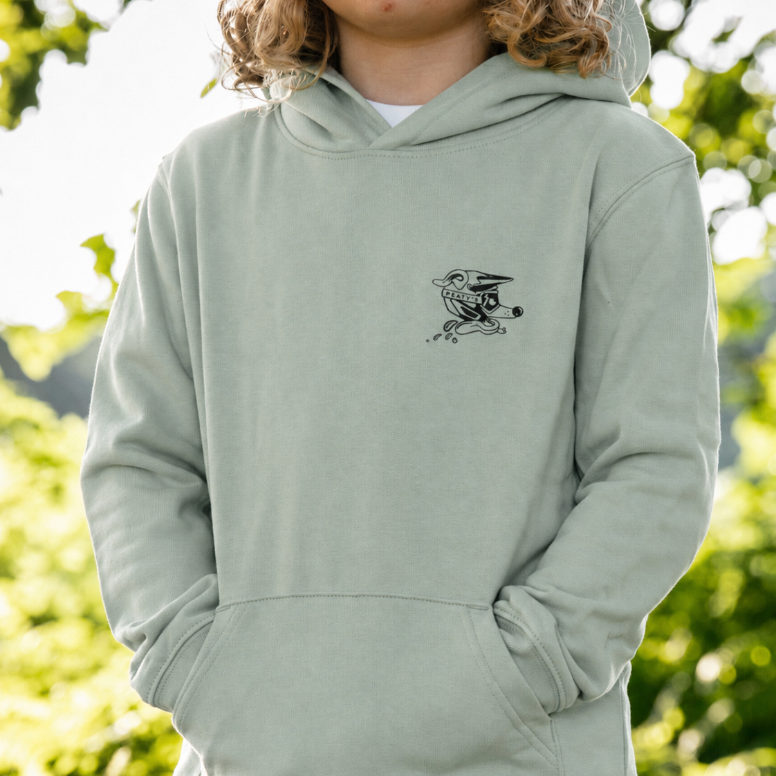 A child wearing a sage green pullover hoodie with a small black bird graphic on the left chest, photographed outdoors against a blurred green foliage background.