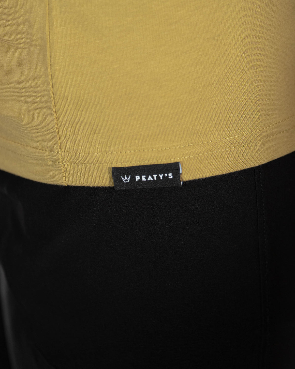 Close-up of the Peaty's woven hem tag on the Olive Dusk to Dawn T-shirt