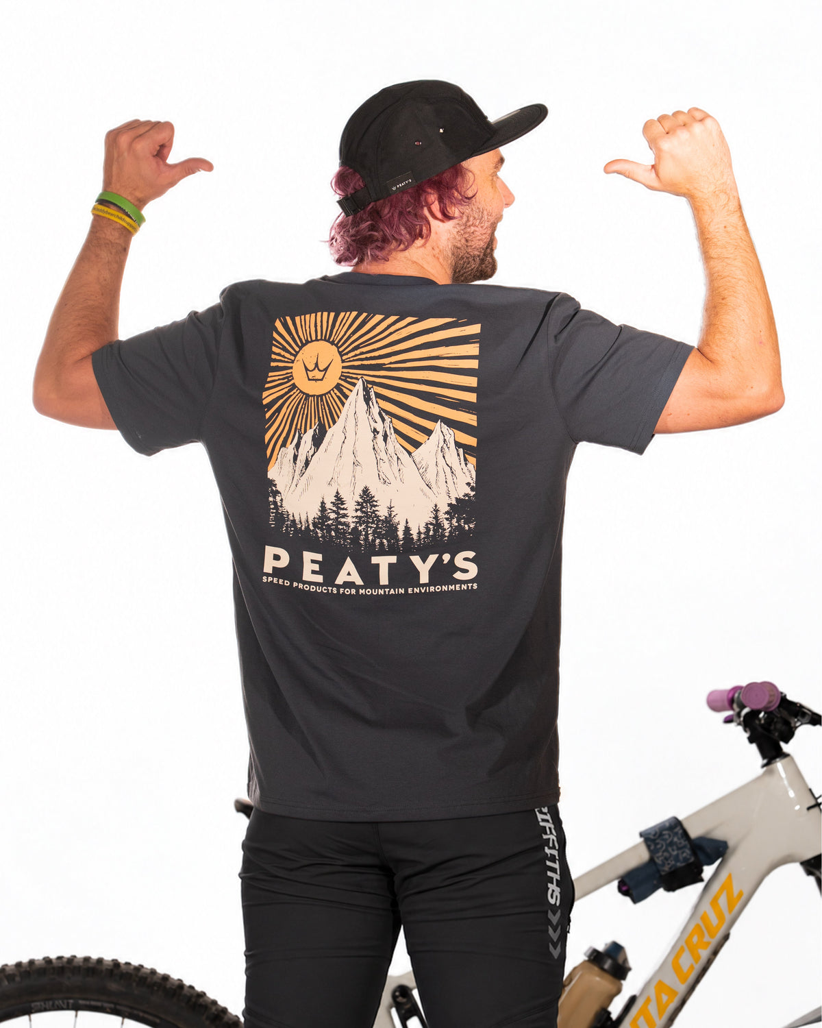Rider wearing dark navy Peaty's Mountain T-shirt beside a mountain bike, with back graphic